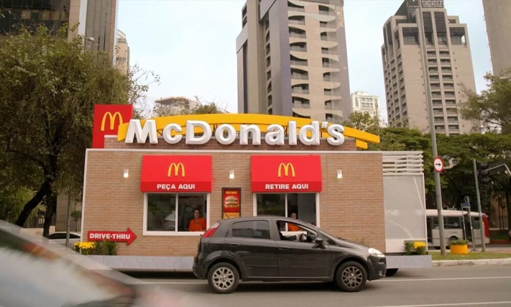 McDonald’s inaugura primeiro drive-thru na Serra em Colina de Laranjeiras Drive-thru do McDonald’s com carro no atendimento da janela “Peça Aqui”, em área urbana com prédios ao fundo.