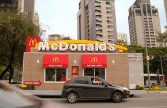 Drive-thru do McDonald’s com carro no atendimento da janela “Peça Aqui”, em área urbana com prédios ao fundo.