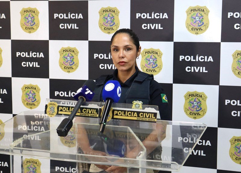Policial civil mulher em pé à frente de microfones de emissoras de TV durante coletiva de imprensa sobre o caso de tentativa de feminicídio Serra ES, com o painel da Polícia Civil ao fundo.