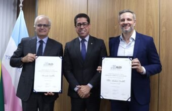 Na imagem, Ricardo Botelho e Fabio Bertollo seguram os quadros com o titulo cidadao espirito santense ALES, posando ao lado do presidente da Assembleia, Marcelo Santos, durante a cerimônia oficial.