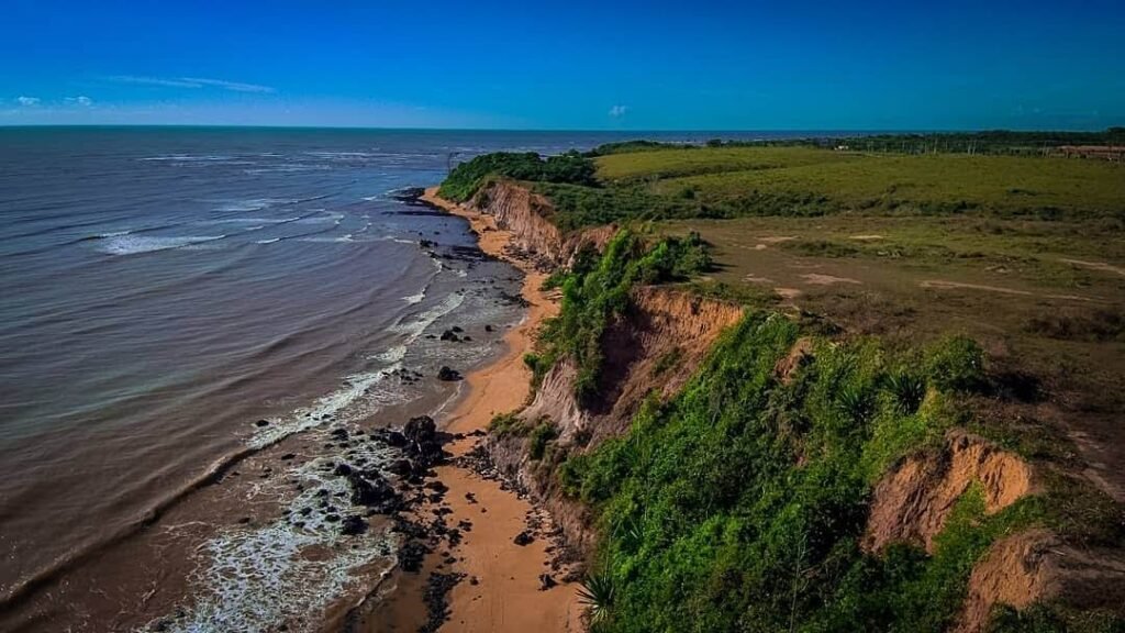 Vista aérea das falésias de Nova Almeida em Serra, Espírito Santo, com vegetação densa no topo, faixa de areia estreita e rochas na base banhadas pelo oceano Atlântico