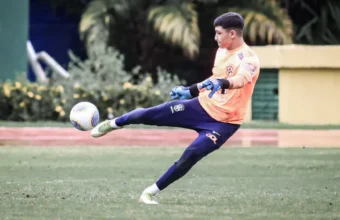 O goleiro capixaba Arthur do Vale chuta uma bola de futebol em um campo gramado, vestindo o uniforme de treino da seleção brasileira composto por camisa laranja de manga longa, calça azul e luvas azuis.