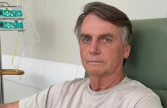 Fotografia do ex-presidente Jair Bolsonaro sentado em uma poltrona clínica de hospital, vestindo camiseta clara, ao lado de equipamentos de infusão venosa. A imagem de sua internação recente ilustra as dúvidas da população sobre o que é broncopneumonia sintomas e a gravidade dessa infecção respiratória.