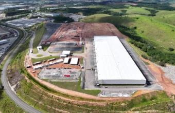 Vista aérea das obras do novo condominio logistico Cariacica ES, mostrando um grande galpão de teto branco já construído ao lado de uma vasta área de terraplenagem preparada para expansão, com uma rodovia contornando o terreno e montanhas verdes ao fundo.