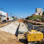 Canteiro de obras em horário de pico na Grande Vitória com gerador CAT e escavação profunda ocupando pistas centrais.