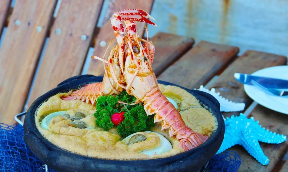 Festival em Manguinhos na Semana Santa tem pratos a partir de R$ 25 Prato de frutos do mar servido em uma tradicional panela de barro preta, decorado com dois lagostins inteiros, salsa e pimenta biquinho. A panela está sobre uma superfície de madeira rústica, ao lado de uma rede de pesca azul e uma estrela do mar decorativa, ilustrando as opções do festival gastronômico Manguinhos Serra.