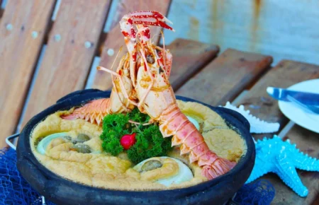 Prato de frutos do mar servido em uma tradicional panela de barro preta, decorado com dois lagostins inteiros, salsa e pimenta biquinho. A panela está sobre uma superfície de madeira rústica, ao lado de uma rede de pesca azul e uma estrela do mar decorativa, ilustrando as opções do festival gastronômico Manguinhos Serra.