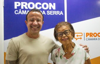 Fotografia do presidente da Câmara, Dr. William Miranda, sorrindo e abraçando a aposentada Clemilda Muniz, que usa óculos e blusa florida. Ao fundo, destaca-se o painel de atendimento do Procon da Câmara da Serra em cores azul, branco e laranja.