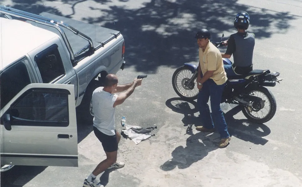 Fotografia diurna da reconstituição de um crime na rua. Um homem de camisa branca e bermuda escura aponta uma arma de fogo simulada para outro homem de camisa amarela e calça jeans, enquanto um terceiro indivíduo aguarda em uma motocicleta azul. Uma caminhonete prata aparece à esquerda com a porta aberta. A encenação policial ilustra o caso que culminou na prisão ex-juiz Alexandre Martins ES.